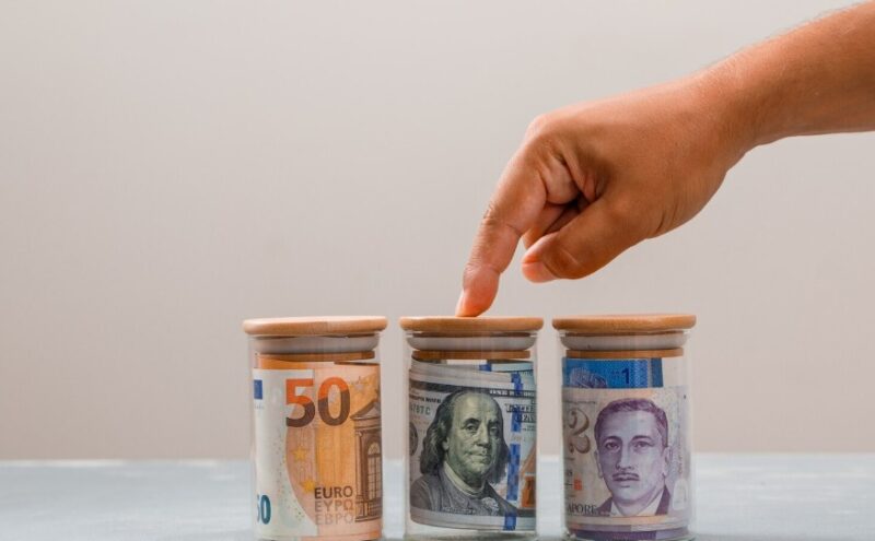 Ruka dodiruje jednu od tri staklene kasice sa novčanicama evra, američkih dolara i drugih stranih valuta