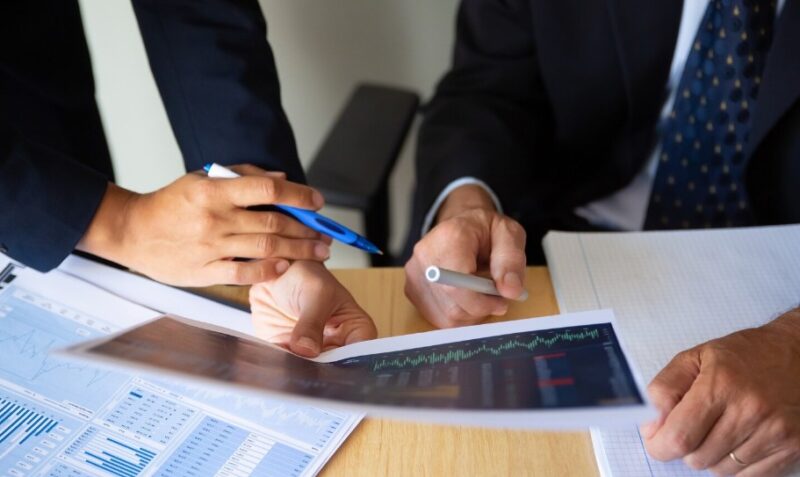 Dva finansijska stručnjaka u poslovnim odelima analiziraju berzanske grafikone i finansijske dokumente za radnim stolom