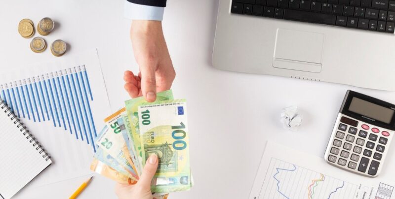 Dve osobe razmenjuju novčanice evra iznad radnog stola sa laptopom, kalkulatorom i finansijskim grafikonima